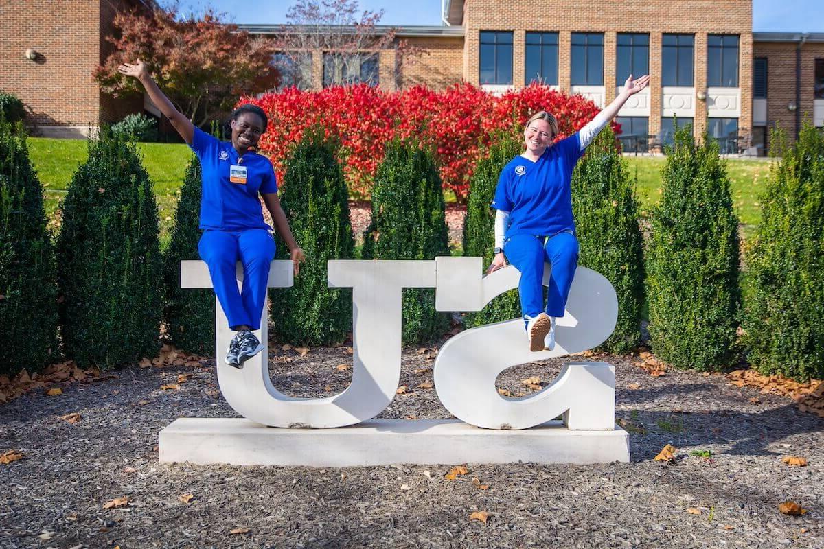 Two Shenandoah nursing students, in blue scrubs, 秋天坐在全球最大的博彩平台广场上的SU雕像上.